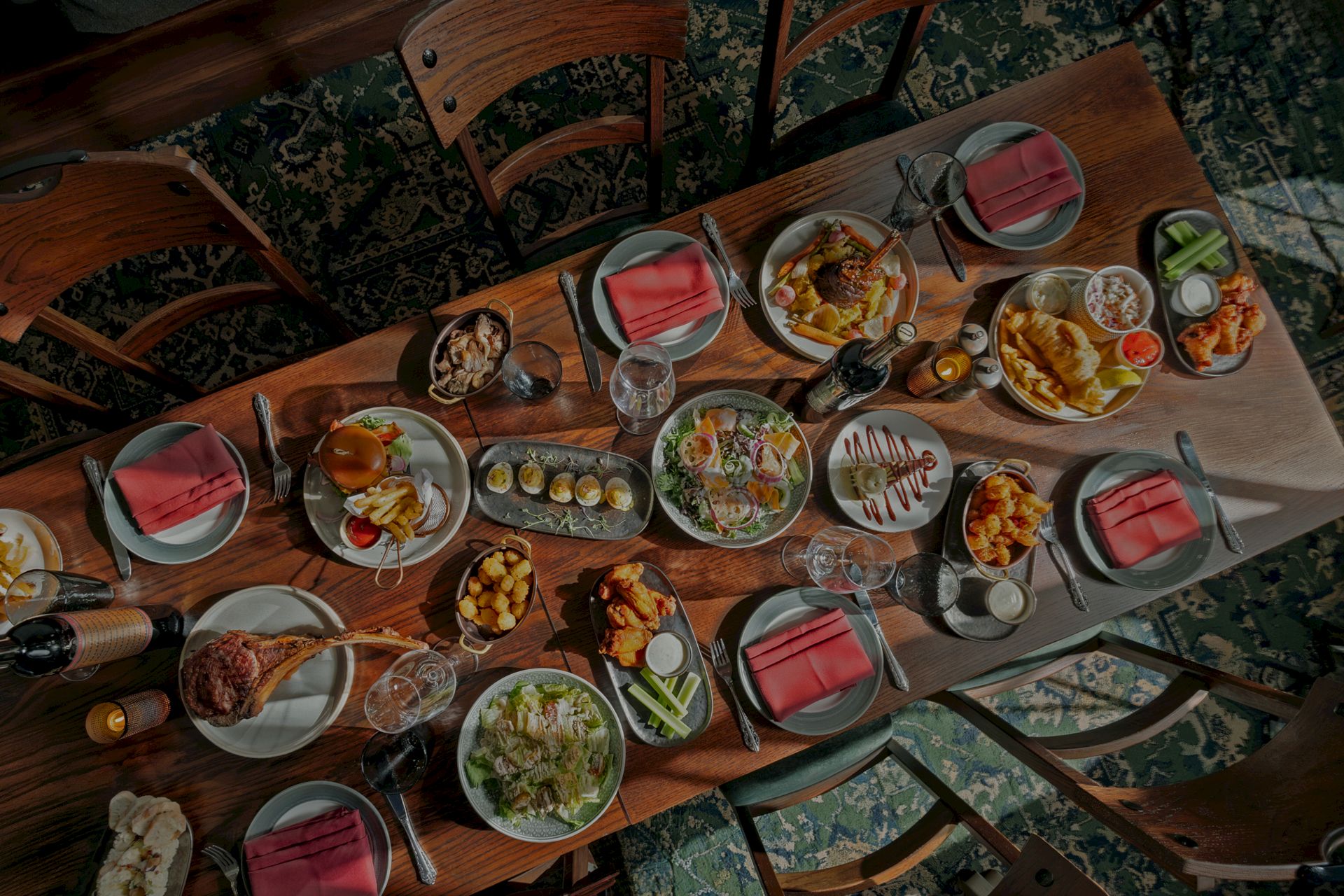 A large dining table is covered with many plated foods, drinks, and red napkins in a cozy restaurant setting, ready for a group meal.