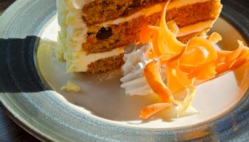 A slice of layered carrot cake with cream cheese frosting, topped with whipped cream and a carrot-flower garnish on the plate.