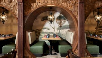 A cozy, rustic restaurant booth with curved wooden partitions, green and cream seating, warm lighting, and wall murals behind the tables.
