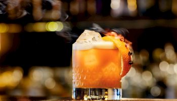A short, stylish cocktail with an orange hue in a textured glass, garnished with a citrus twist, on a dimly lit bar backdrop.