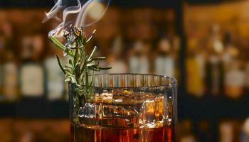 A smoking rosemary garnish over a glass of amber whiskey with a citrus twist on the side, set against a dim bar backdrop.