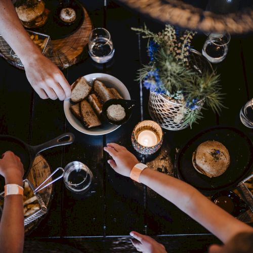 A top-down dinner scene: hands reach for bread and dips, small plates, candles, glasses, and a centerpiece with blue flowers on a dark table.