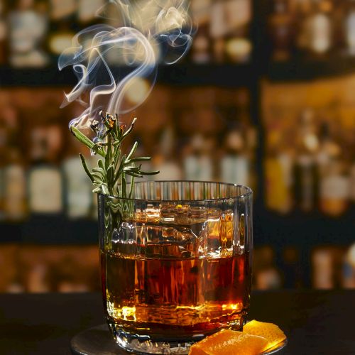 A smoked rosemary whiskey on the rocks with an orange garnish, set against a blurred bar backdrop.