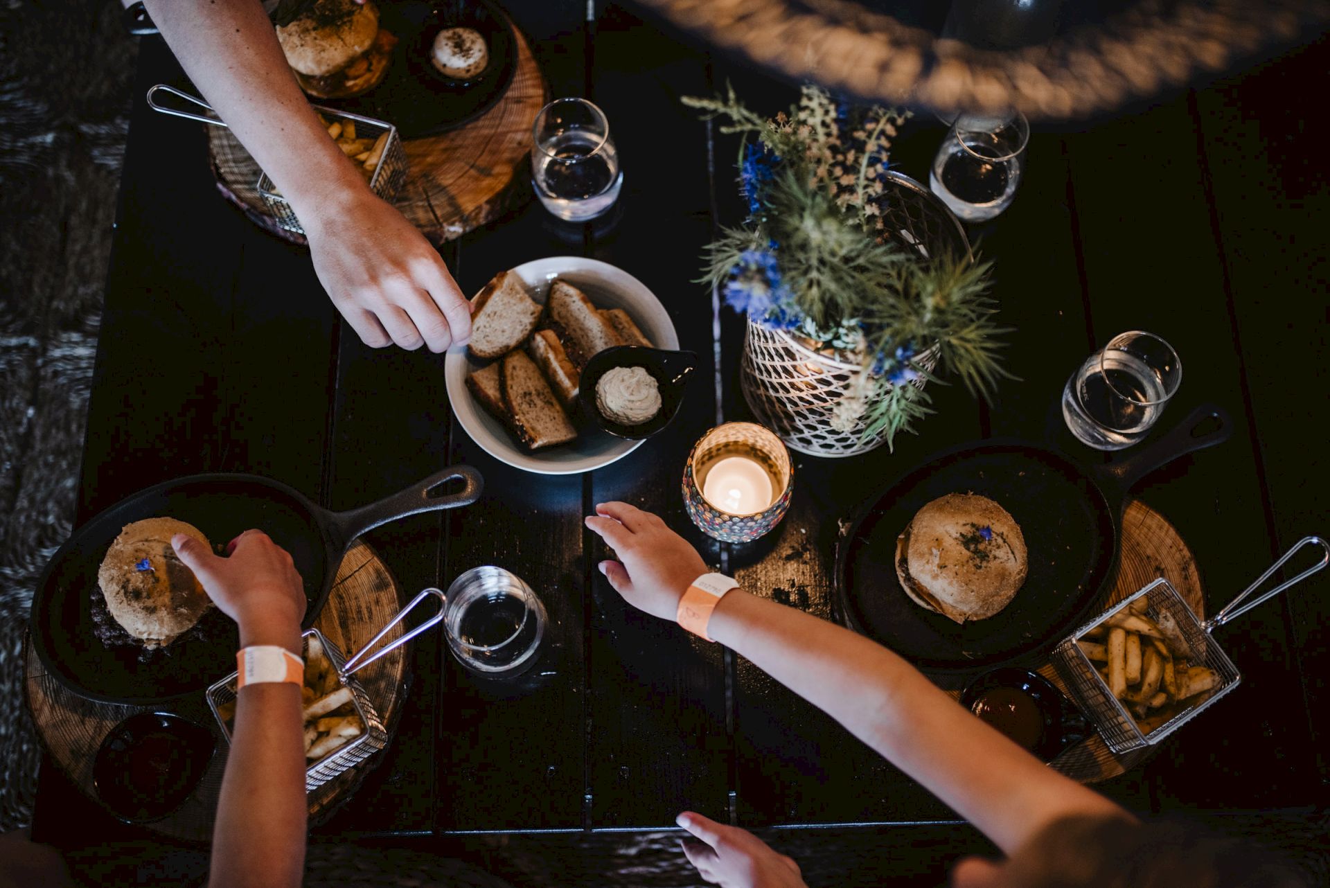 People sharing a dark table meal: hands reach for bread, bowls of soup or dips, candles, glasses, and a flower centerpiece.