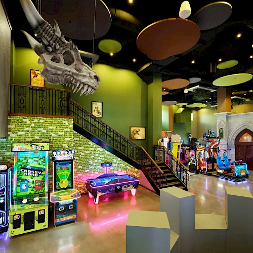 Arcade room with neon games, claw machines, a glowing pool table, and a staircase leading to a mezzanine in a colorful, playful arcade.
