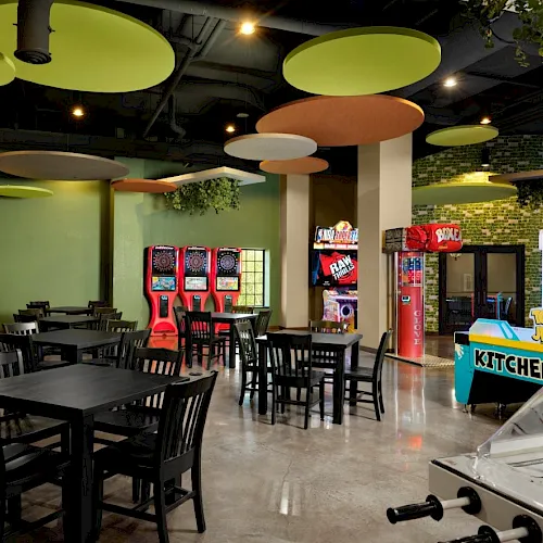 A spacious arcade and dining area with black tables and chairs, arcade games, and a foosball table near a kitchen-themed gaming zone, lively and colorful.