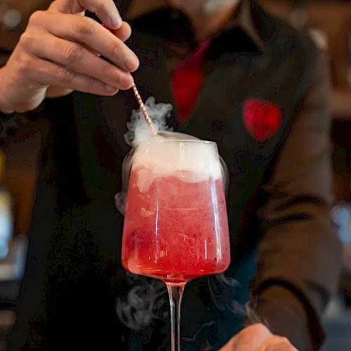 A bartender is pouring a red cocktail into a glass, with smoke rising from the drink.